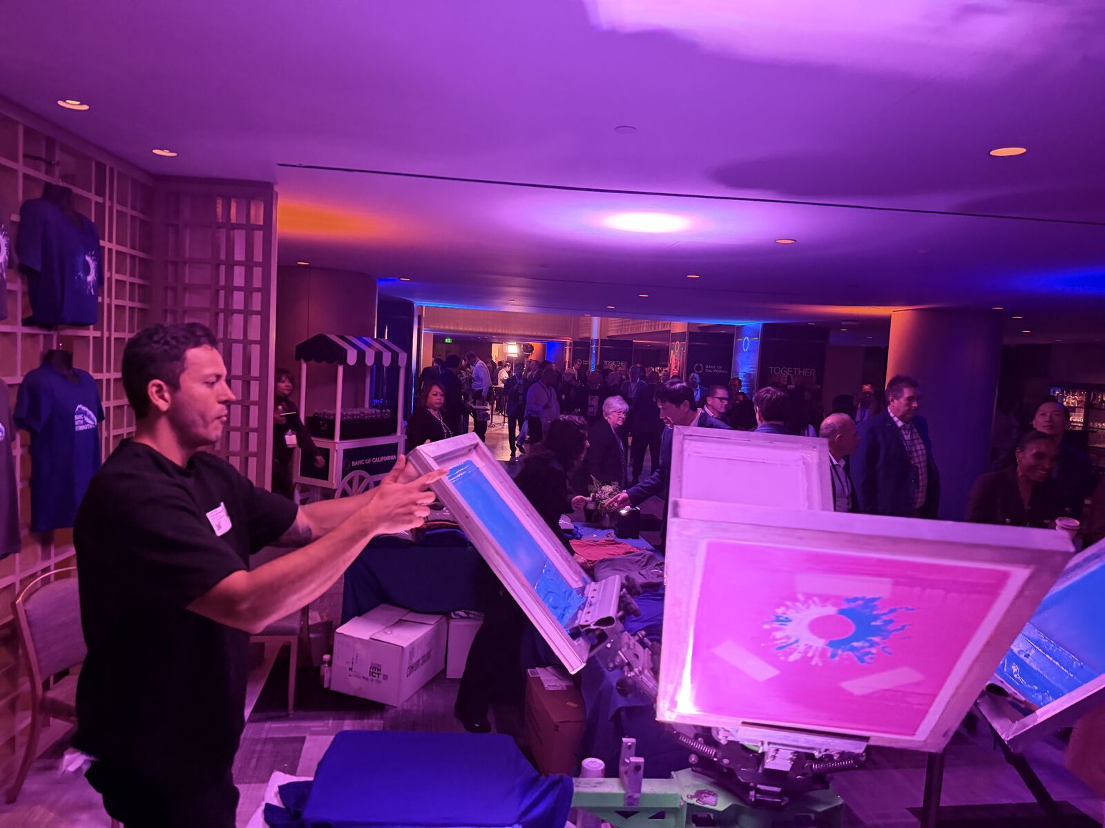 Event booth operator demonstrating a screen printing press with a crowd of attendees gathered around under colorful stage lighting